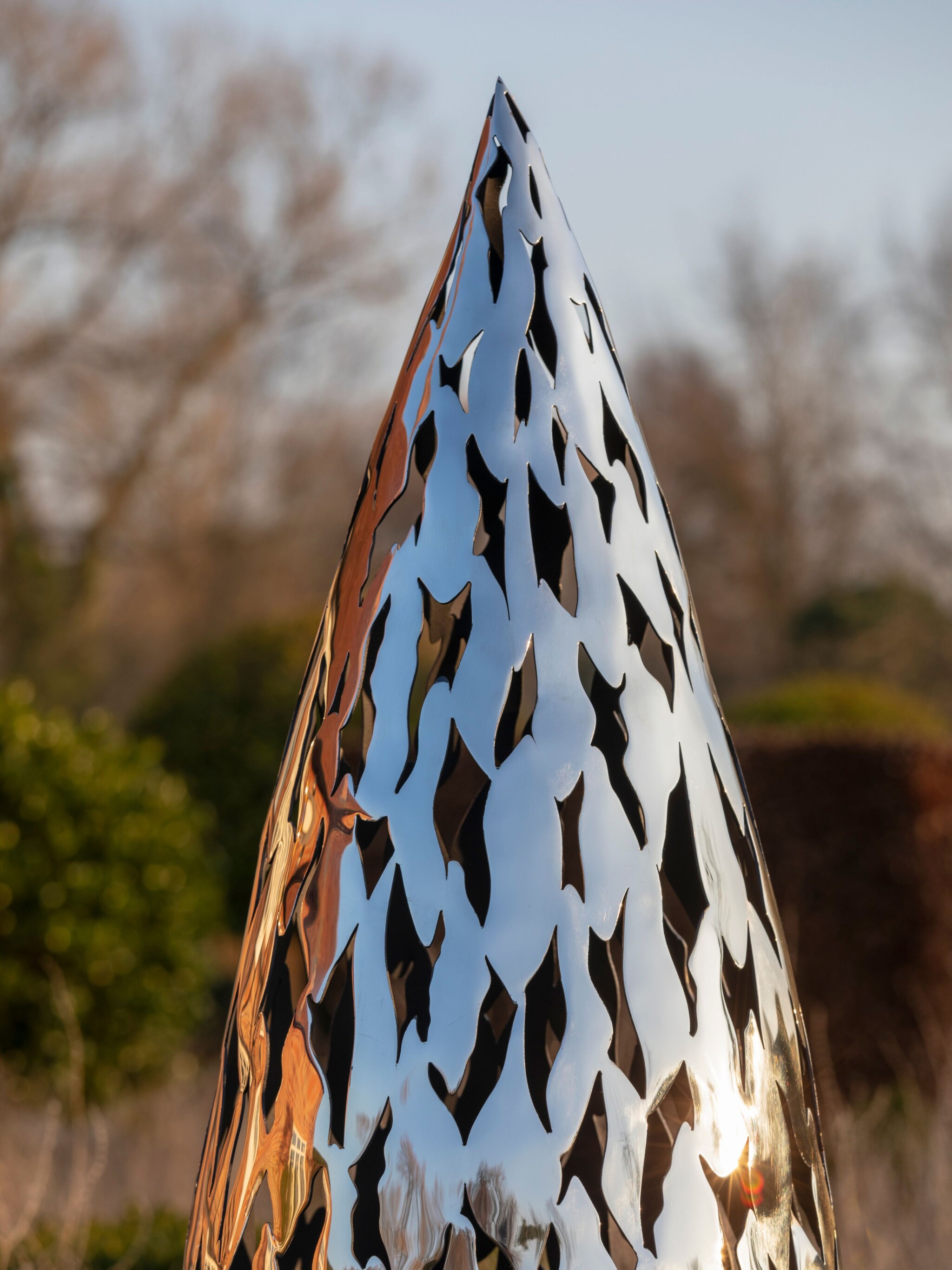 A quill-shaped, shiny stainless steel contemporary sculpture with irregular cutouts set among trees and bushes outdoors.