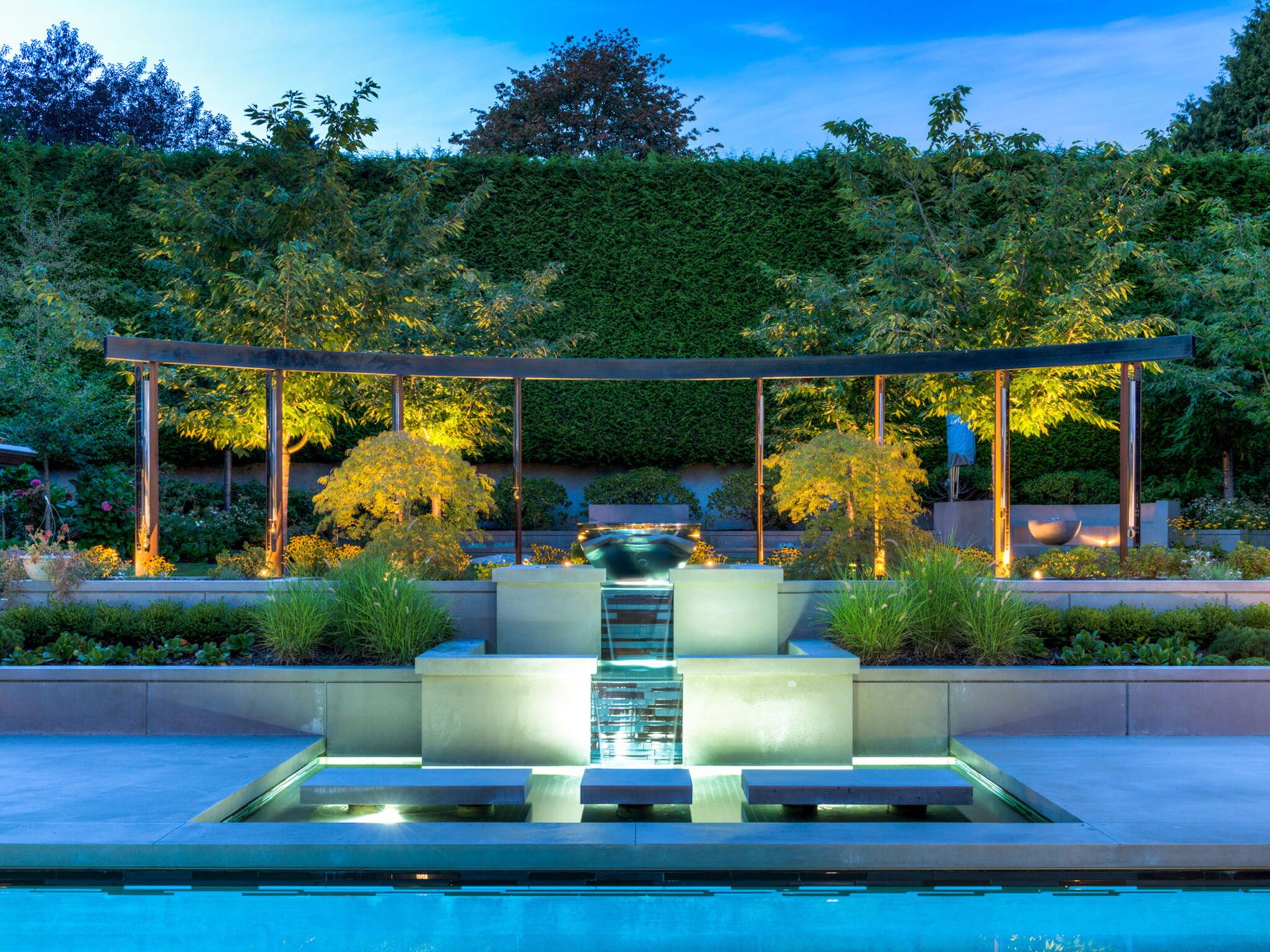 Outdoor lights illuminate a swimming pool and formal garden, which has a giant wind chime sculpture as a centrepiece