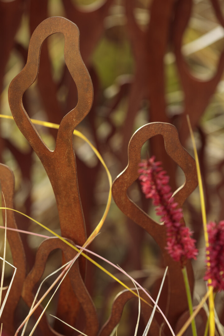 Rust-colored abstract metal shapes among grasses and red flowers, with a blurred natural background.