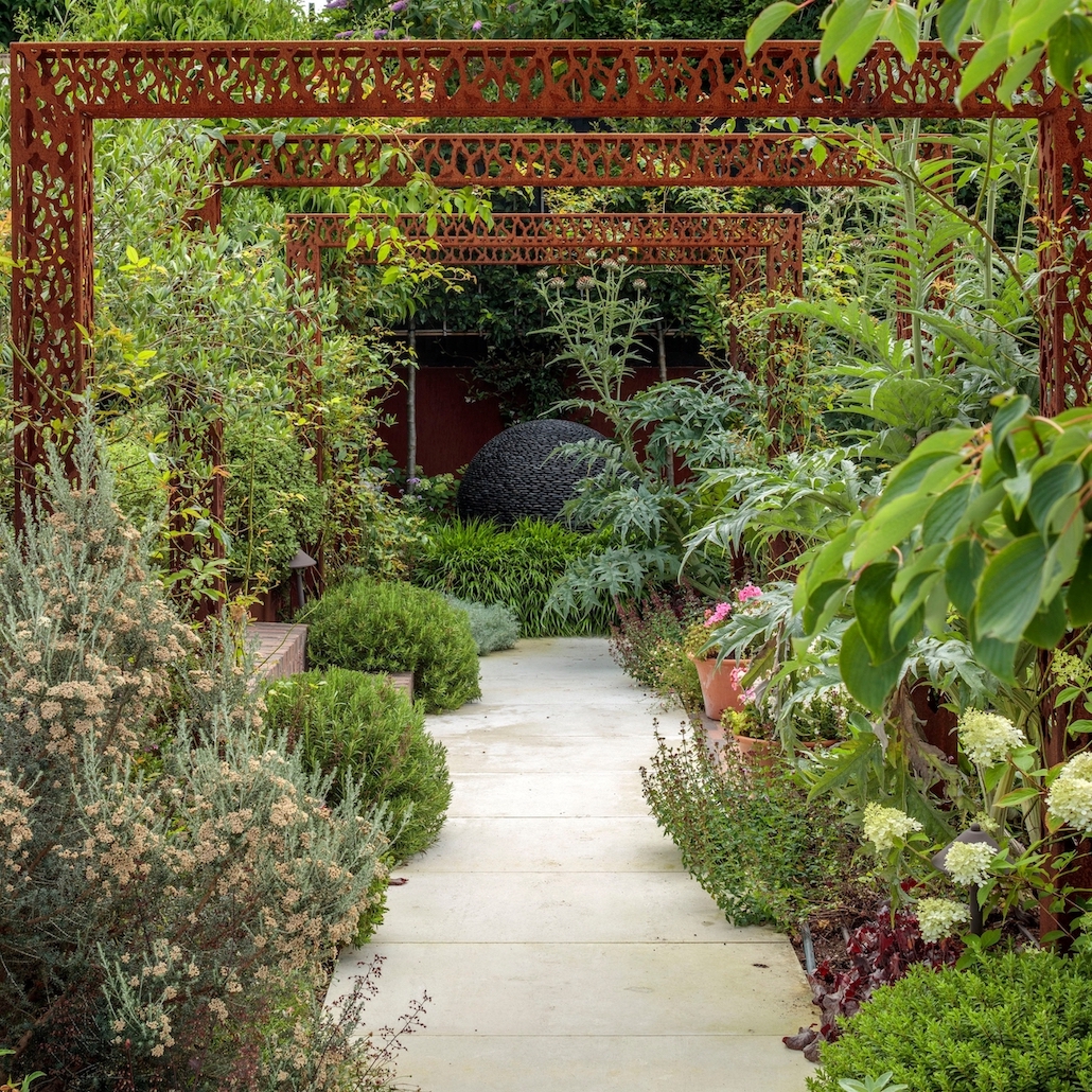 a walkway with plants and a metal arch leading to a focal point of a large, round, pebble sculpture