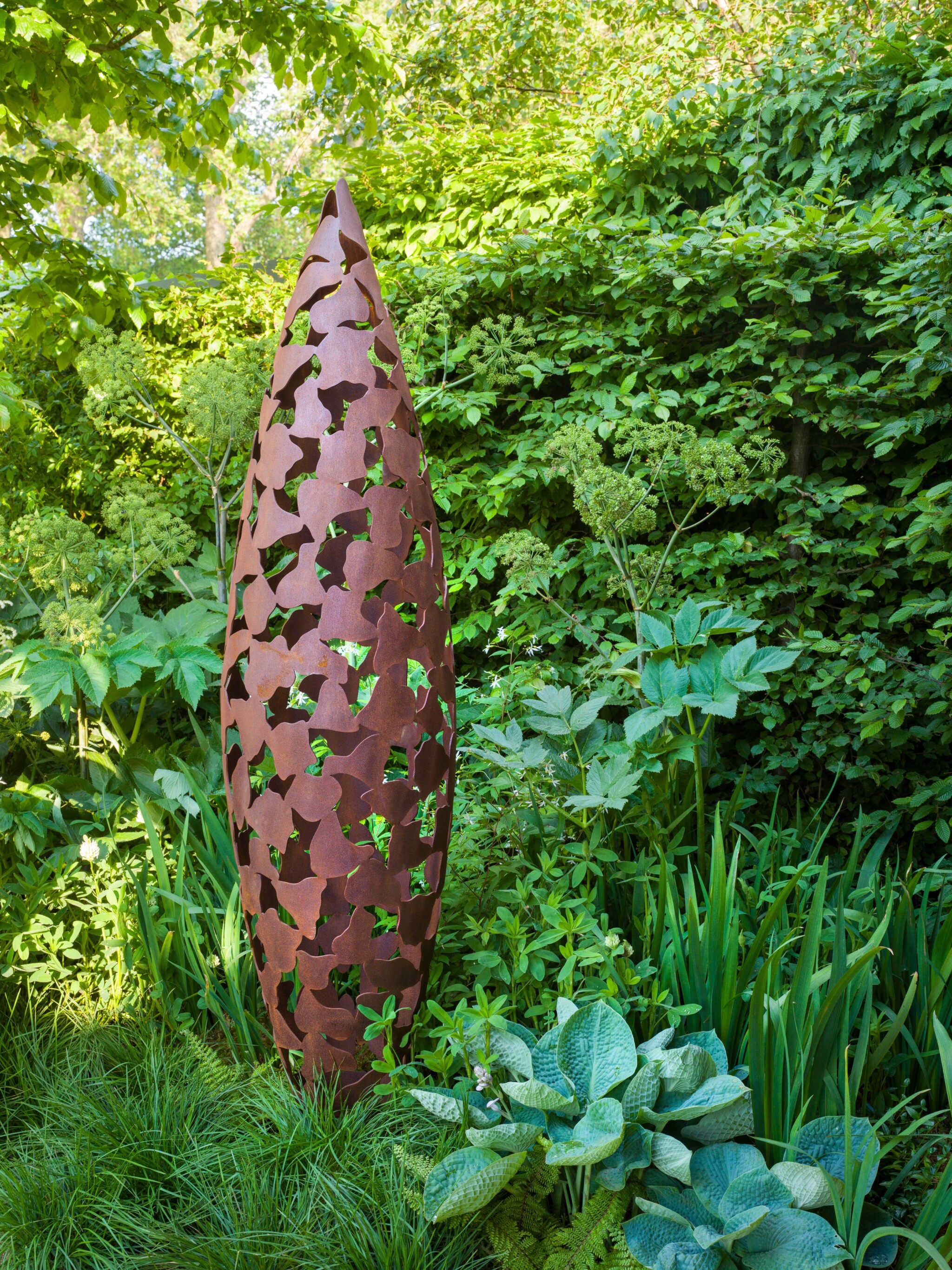 Tall, oval quill oxidised steel contemporary sculpture with cut-out leaves, surrounded by lush green plants and foliage.