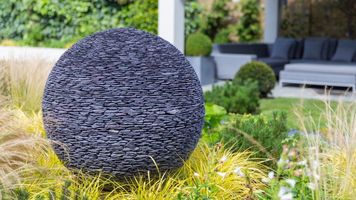 Large round sculpture like a dark planet, made of stacked black smooth stones in a landscaped garden with grass.