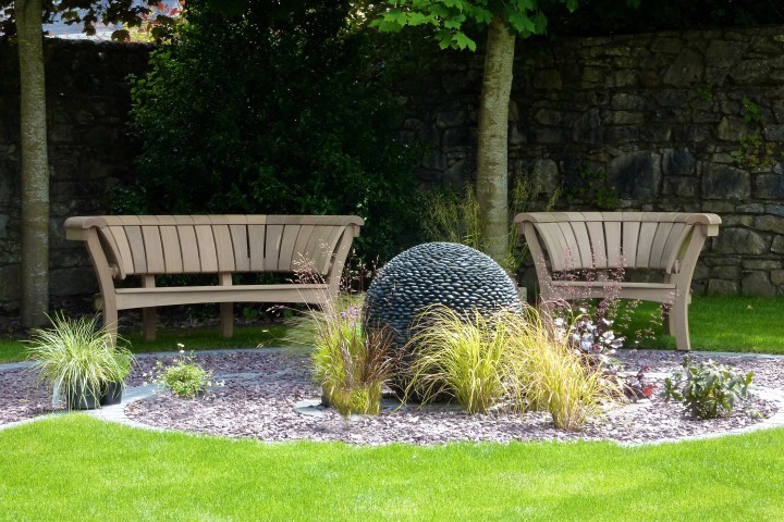 Two wooden benches face a dark planet stone contemporary sculpture in a landscaped garden with grass and plants.