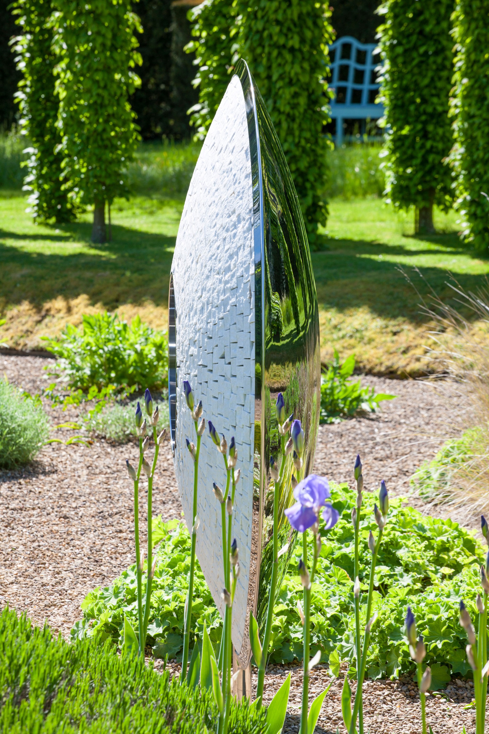 A tall, modern stainless steel sculpture stands among green plants and purple irises in a sunny garden.
