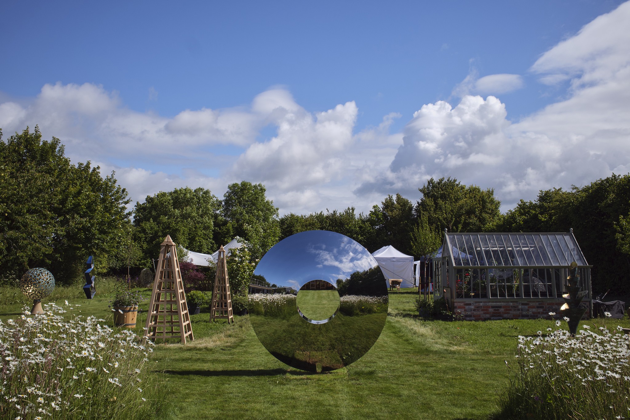 A torus stainless steel contemporary abstract sculpture stands in a garden with flowers, trees, a glasshouse, and marquees.