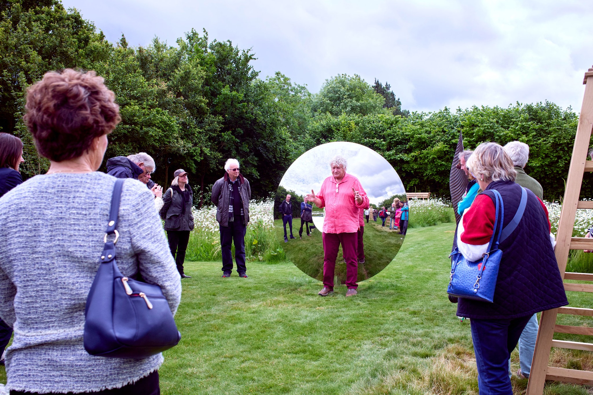 A group of people stand in a grassy field near wildflowers, trees, and a torus stainless steel contemporary sculpture, listening to a person in a pink shirt highlighted by a circular inset effect. The sky is cloudy and most people wear jackets or sweaters.