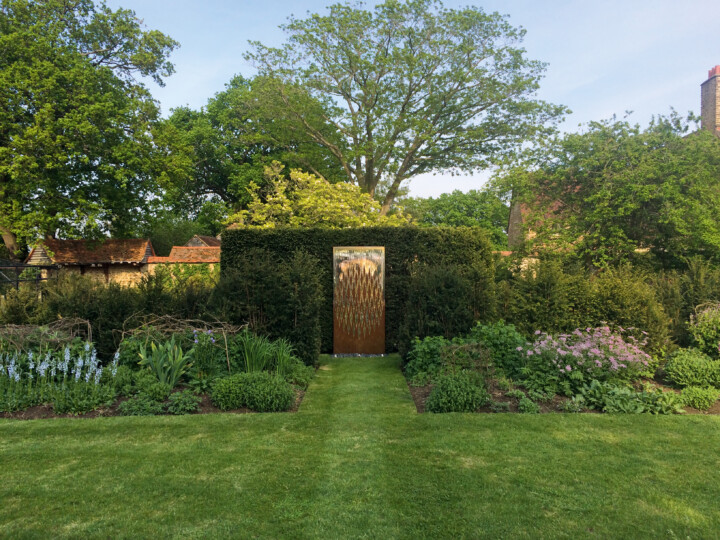 A formal garden with neatly mown grass, lush green hedges, and flower beds. At the centre, an interwoven water wall made from stainless and mild steel stands beside a decorative metallic gate within a tall hedge. Traditional rooftops rise in the background.