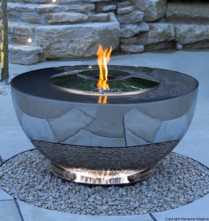 Contemporary fire chalice with a stainless steel surface, flames dancing at the center, and a stone backdrop.