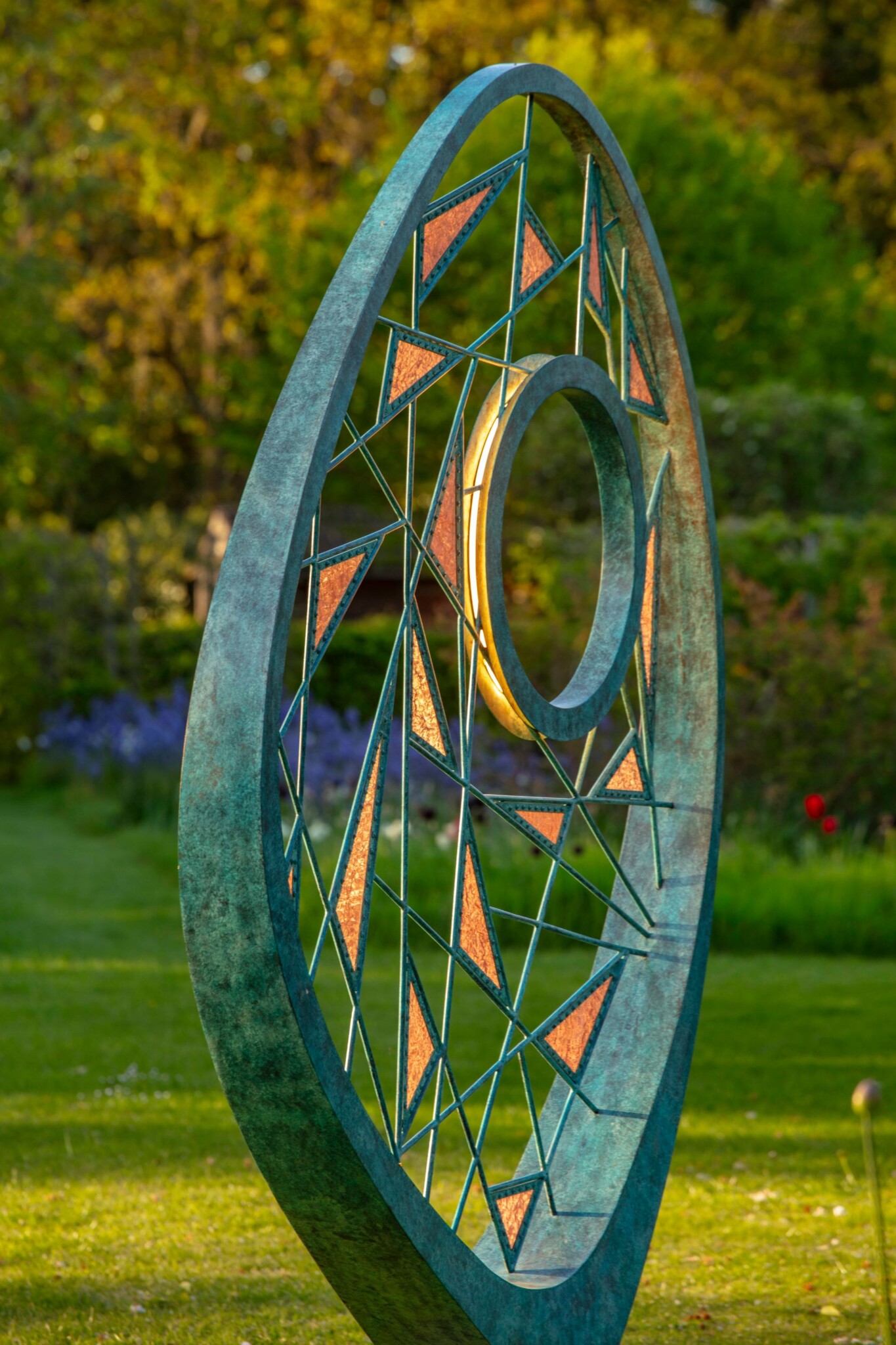 A large metal sculpture made from woven verdigris bronze metal work sits in a formal garden setting, and is illuminated by the glow of evening light.