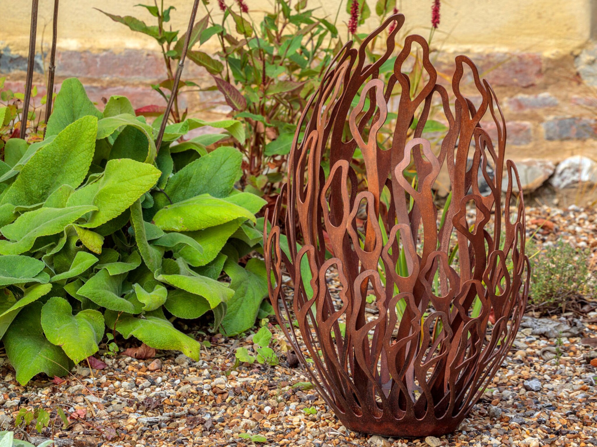 A rust-coloured, metal garden sculpture with flame-like cut-outs stands on a gravel surface next to leafy green plants, with a brick and yellow render wall in the background.