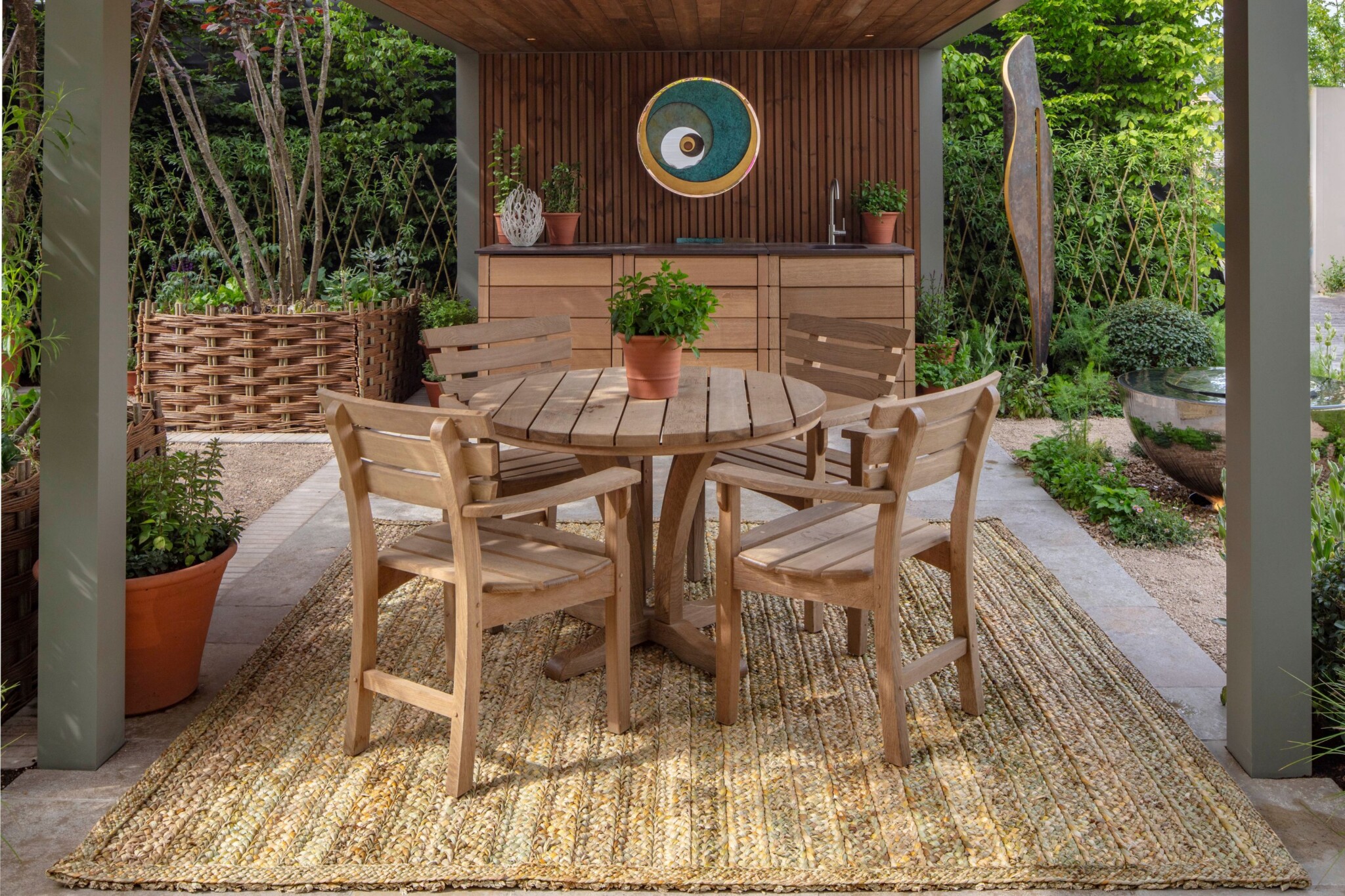 Wooden table and chairs on a woven mat under a structural canopy with a decorative round sculpture on the wall behind