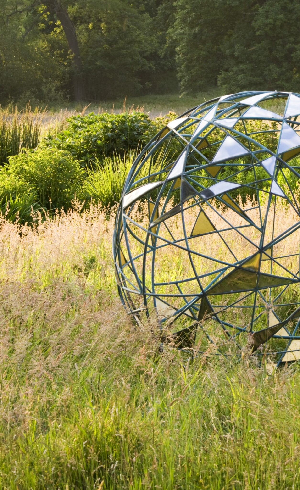 Matrix stainless steel sculpture set in tall grass with green bushes and trees on a sunny day.