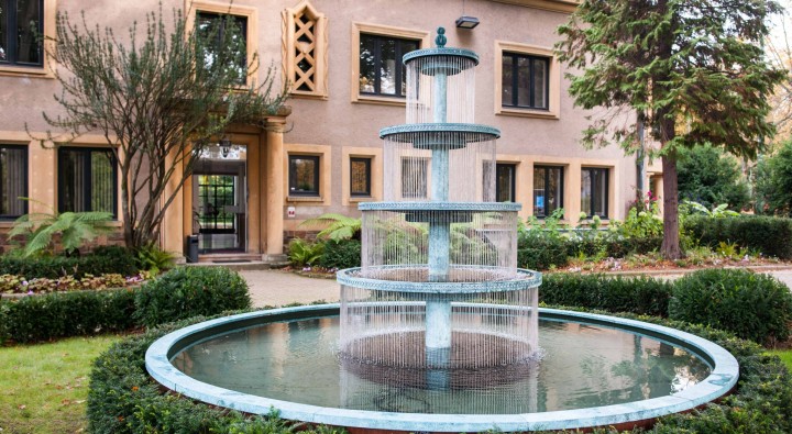 Crucello water fountain cascades in tiers into a circular pool, gracing a garden by a beige building with large windows.
