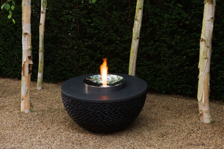A modern dark planet fire table with a burning flame, surrounded by trees on a gravel surface.