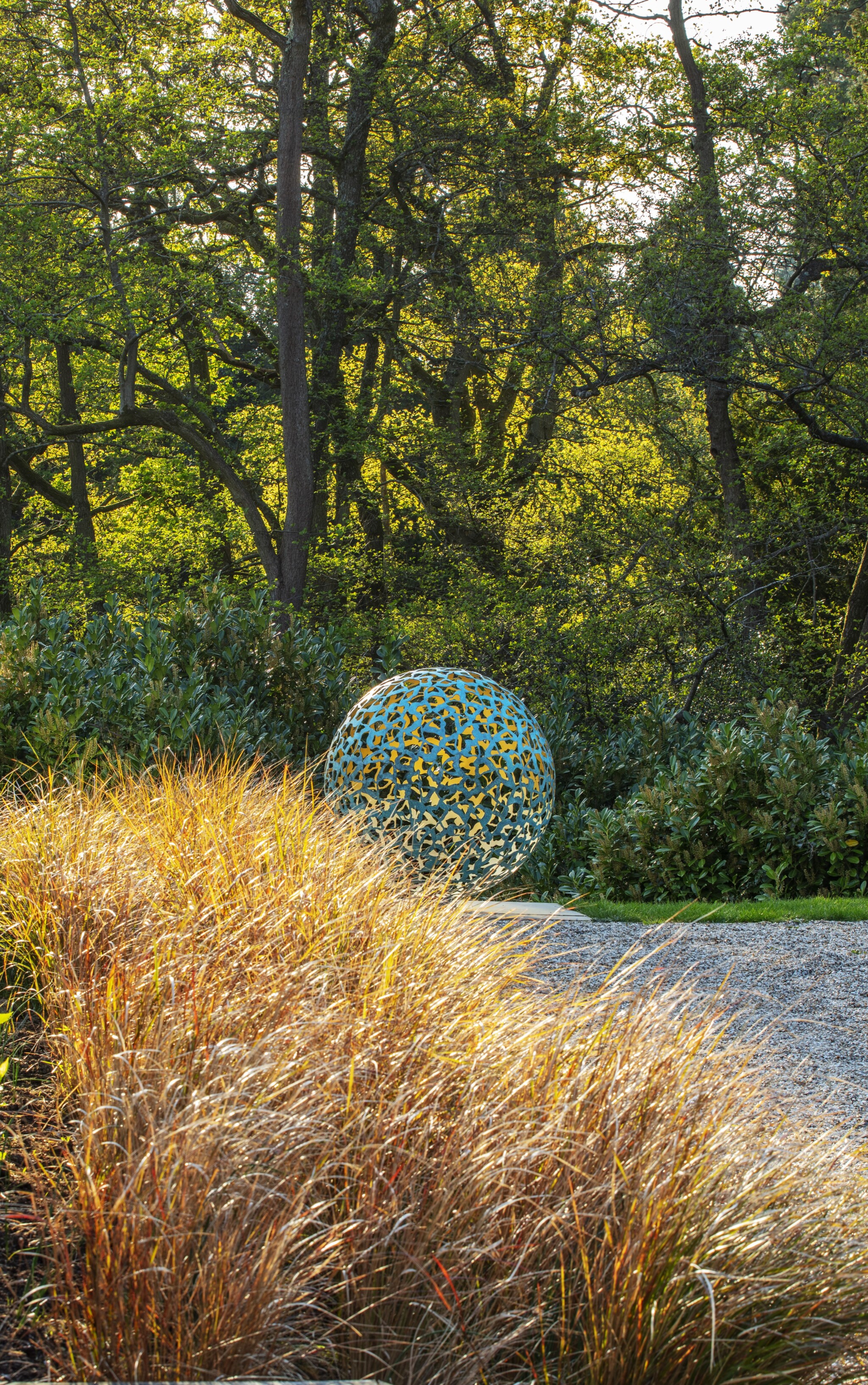 Geminus bronze and gold contemporary abstract sculpture set in a garden with tall grass and lush green trees.