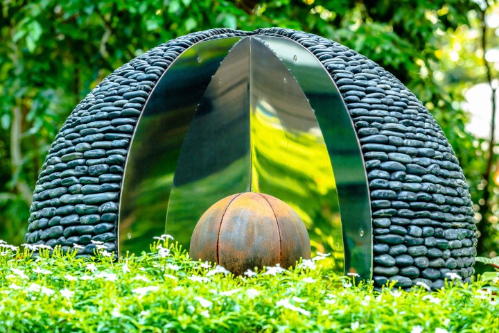 Sculpture with black stones and stainless steel panels encasing a rusted sphere, set in greenery.