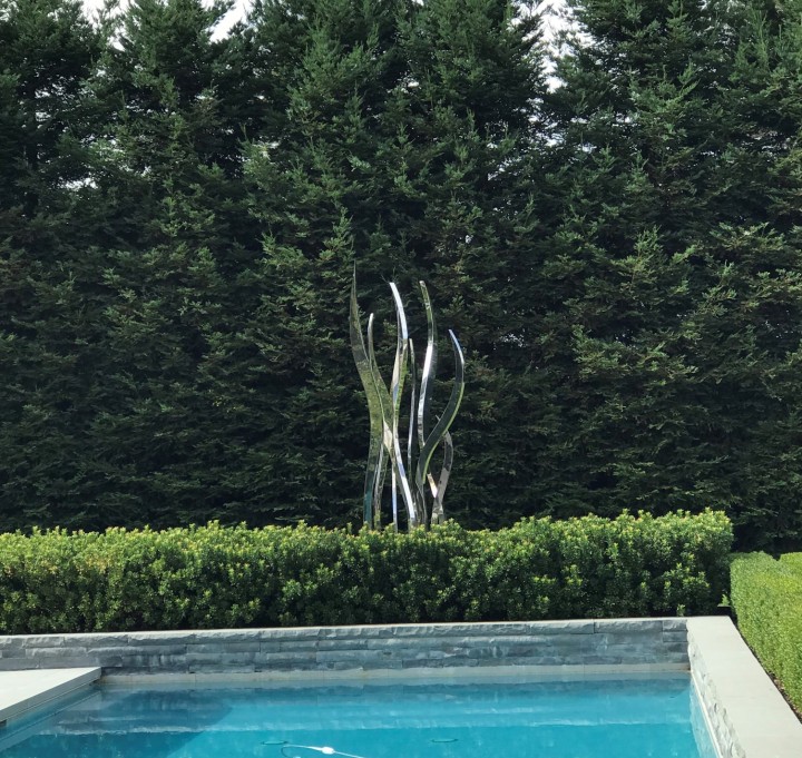 A hydra stainless steel contemporary abstract sculpture rises behind a swimming pool and lush green shrubs, framed by evergreens.