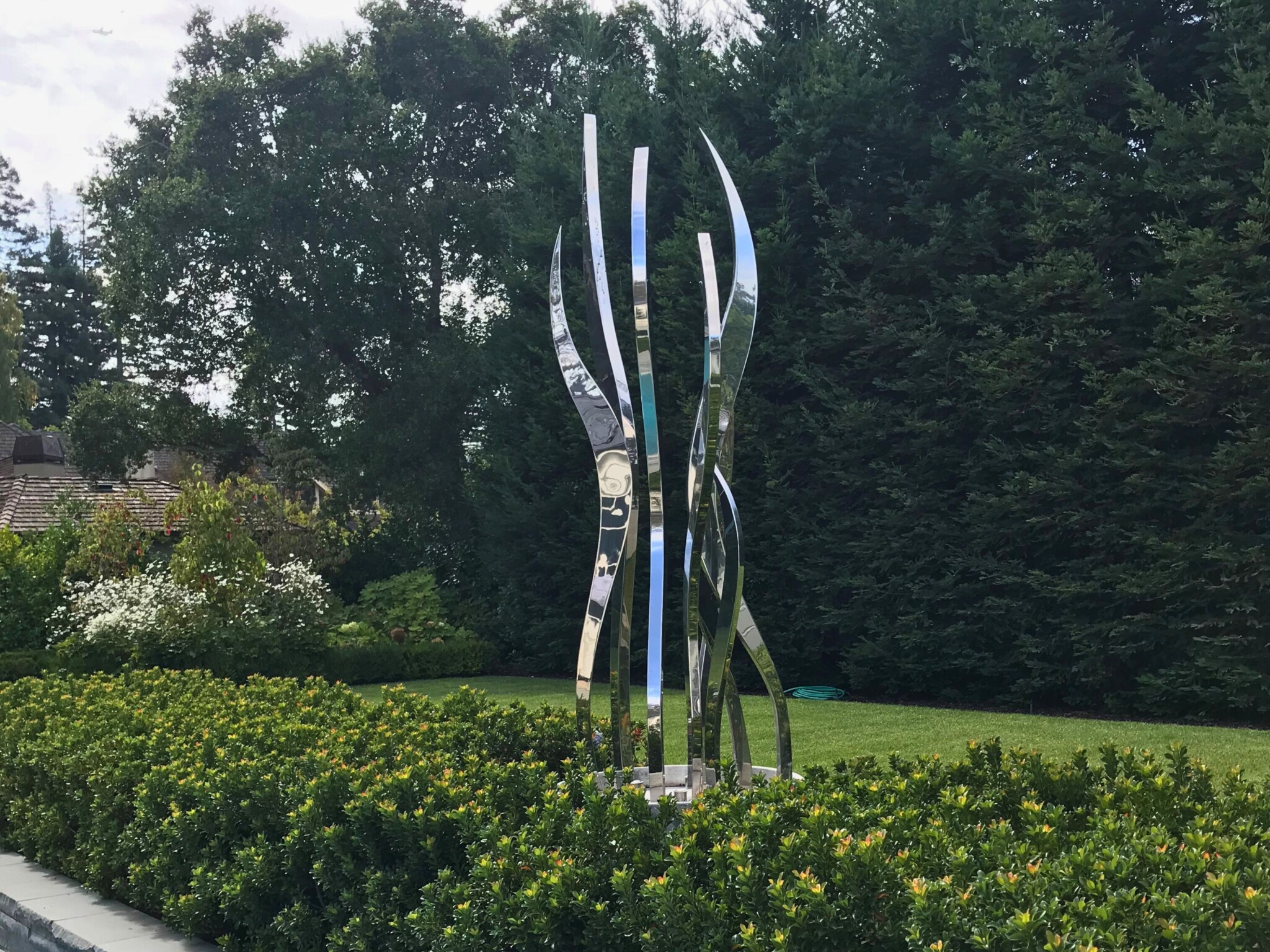 Tall hydra stainless steel contemporary abstract sculpture stands among manicured bushes with trees and lawn behind.