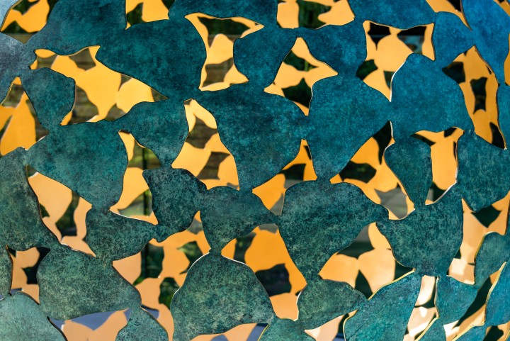 Close-up of Mantle II in bronze and gold leaf, a contemporary abstract sculpture featuring irregular, leaf-like shapes with a weathered teal finish. Warm yellow light shines through the gaps, highlighting the cool metal against the bright background.