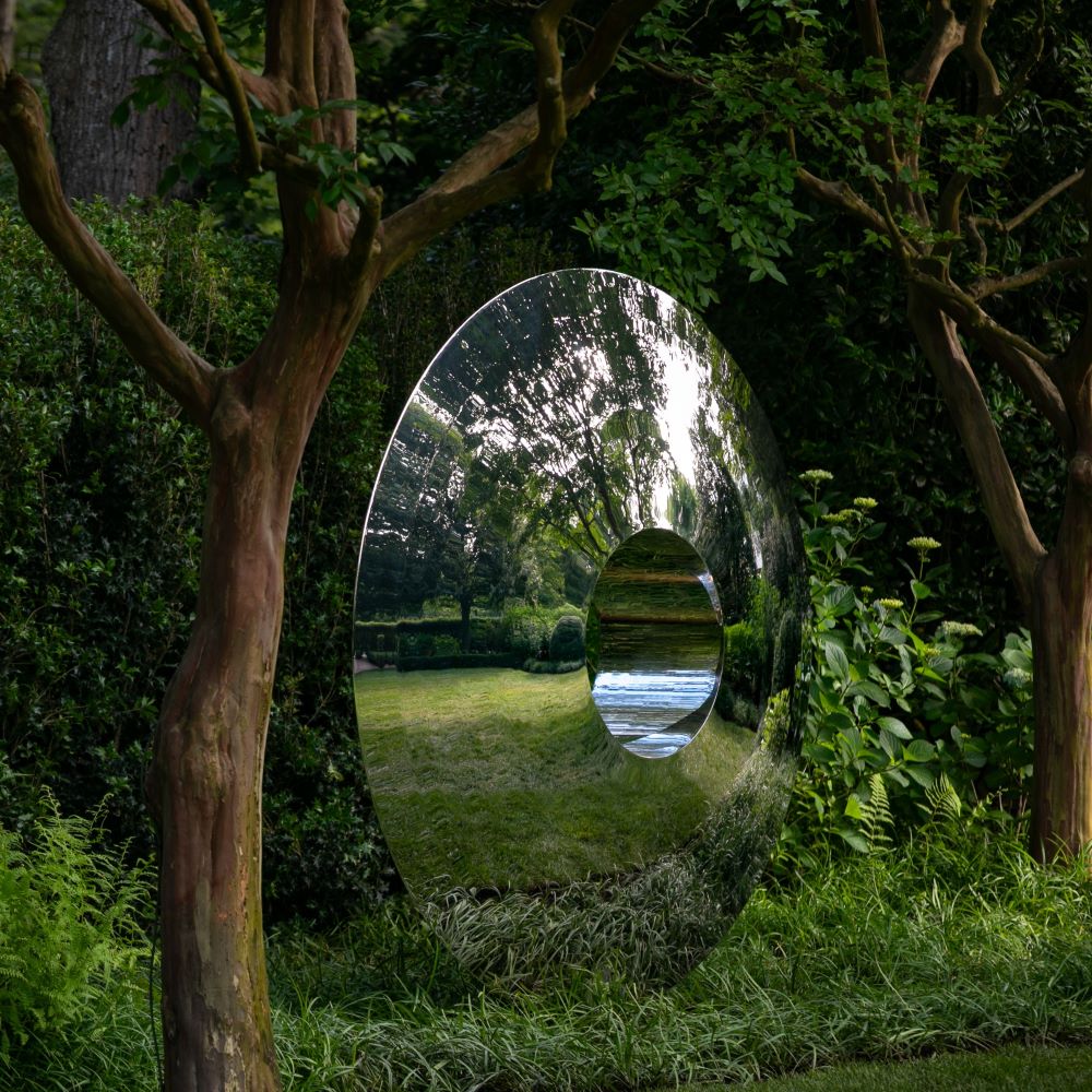 A shiny, circular torus stainless steel contemporary abstract sculpture with a hole in the center stands upright on grass among trees and lush greenery, reflecting the East Hampton garden and New York sky in its mirrored surface.