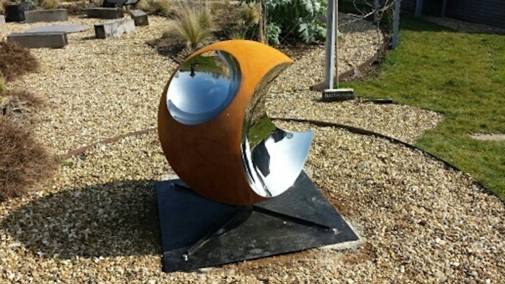 A modern outdoor piece, Bite II is an abstract contemporary sculpture featuring a rusty orange, oxidised steel sphere with a shiny silver “bite” cut away. It stands on a black metal base amid gravel landscaping and green grass.
