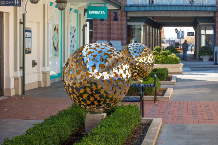 Two large Mantle II in stainless steel and gold leaf contemporary abstract sculptures sit on plinths along a brick pavement lined with greenery in a shopping area, with shopfronts like DAVIDsTEA and signs hanging from awnings behind them.