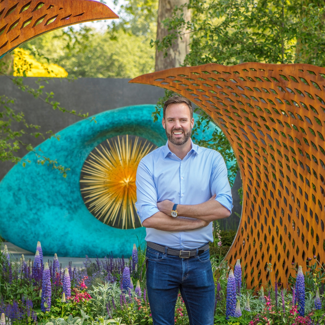 a man standing in front of a large patinated sculpture in a planted garden