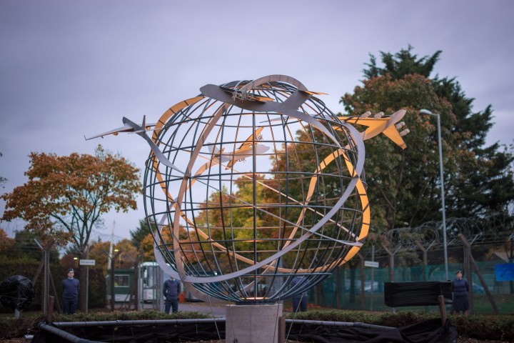 A contemporary abstract sculpture featuring multiple interlocking loops of stainless steel with four of Brize Norton’s key aircraft circles a metal globe, set outdoors among autumn trees, a wire fence, and an overcast sky.