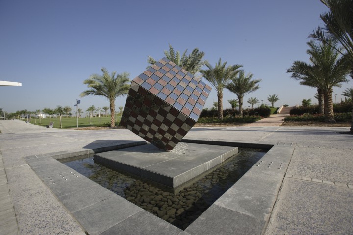 The Cube Contemporary Sculpture at Zabeel Park in Dubai is a large metallic cube with a chequered pattern, balanced on one corner above a shallow rectangular pool, surrounded by palm trees and a landscaped walkway under a clear blue sky.