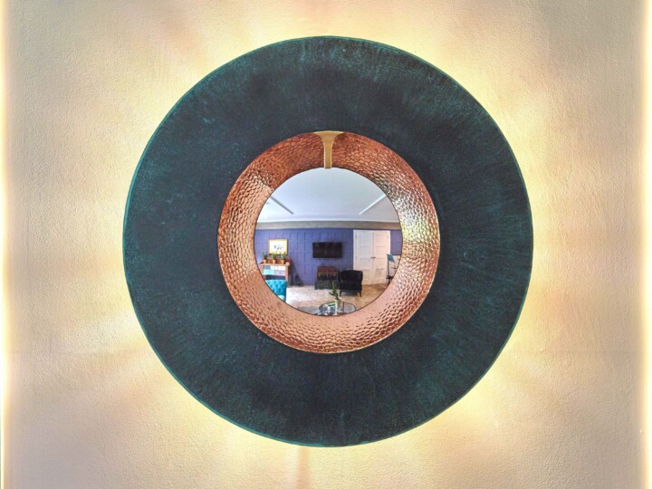 A round mirror with a thick, textured mirage bronze frame hangs on a beige wall. The mirror reflects a cozy room with a couch, TV, table, and blue-paneled wall, while warm ambient lighting radiates softly around the mirror.