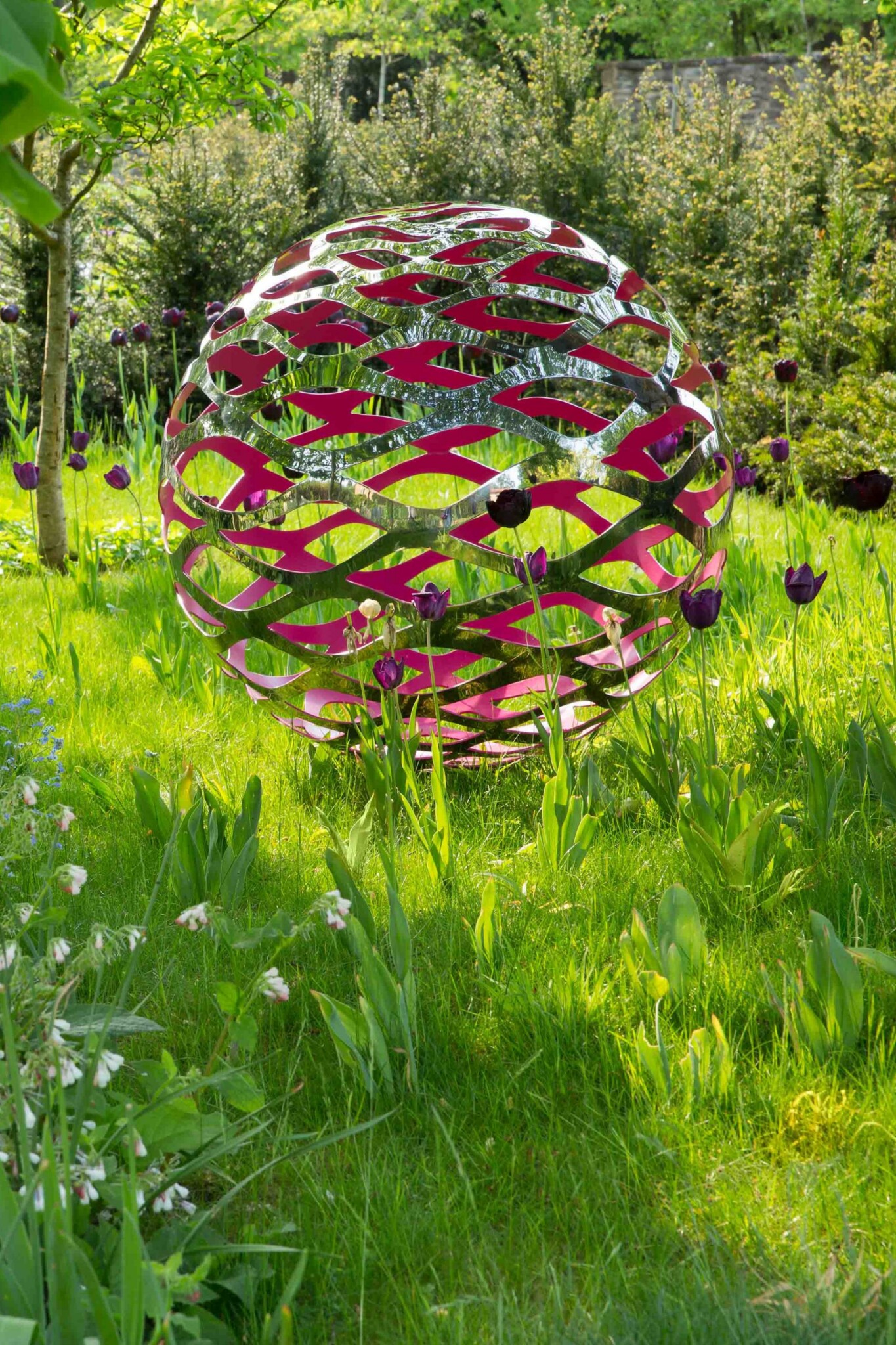 Stainless steel ball sculpture with a painted pink interior and wave-like cut outs, located in a spring garden.