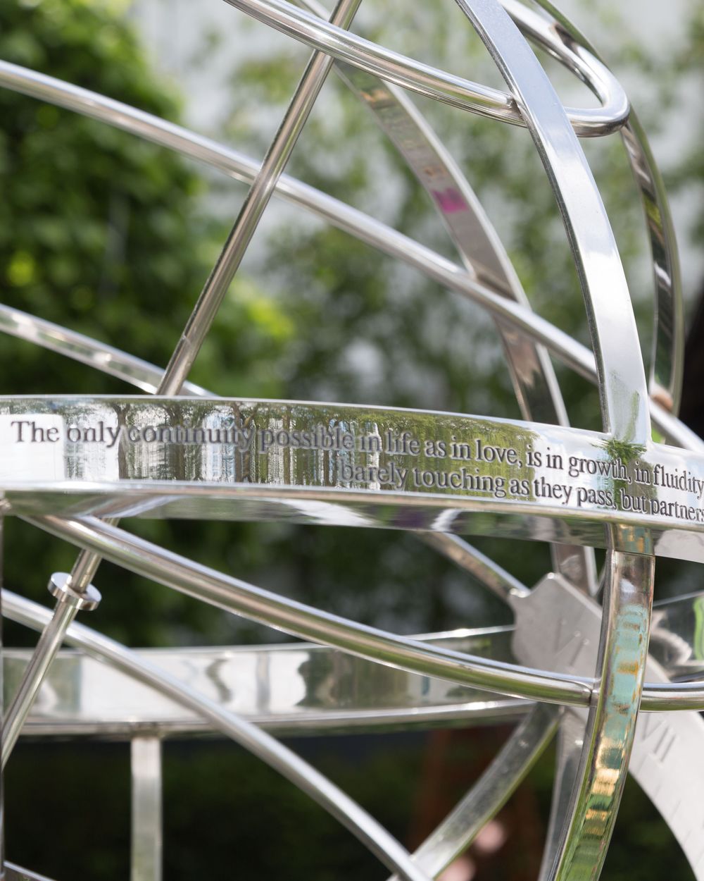 Close-up of a stainless steel sculpture, reminiscent of an armillary sphere, engraved and set outdoors amid greenery.