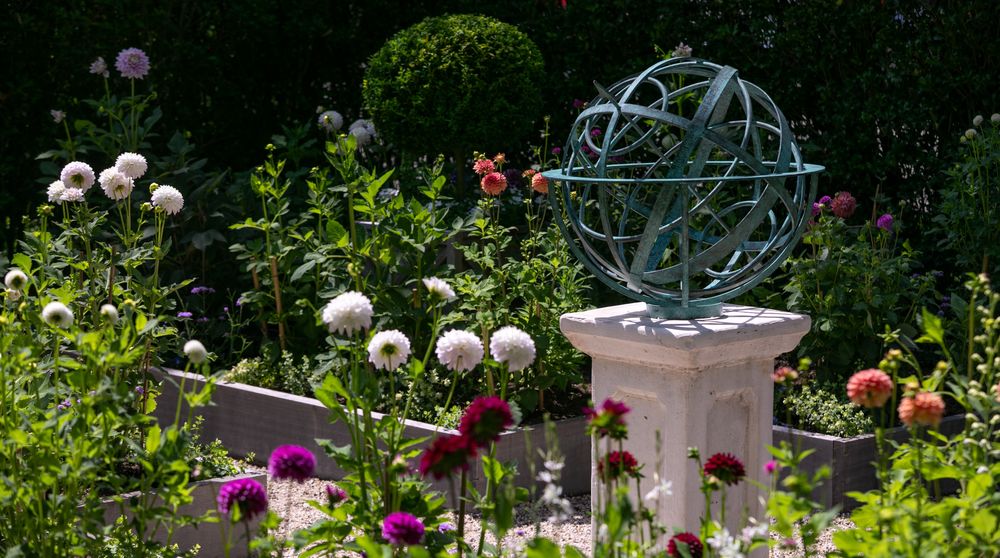 A metal armillary sphere on a pedestal surrounded by blooming flowers in a garden.