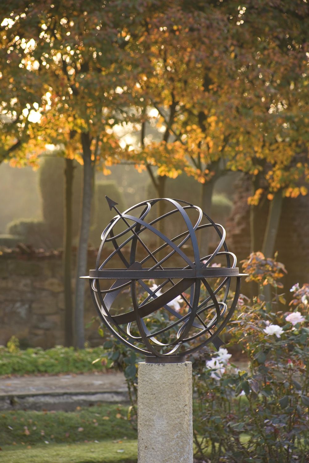 Metal armillary sphere garden sculpture adds elegance on a stone pedestal amid autumn trees.