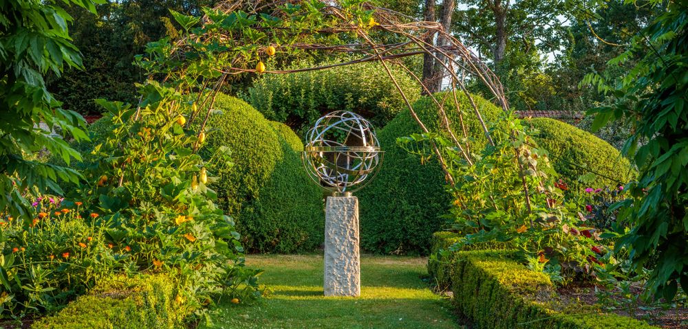 Armillary sphere sculpture on a stone pedestal under a leafy archway in a manicured garden.