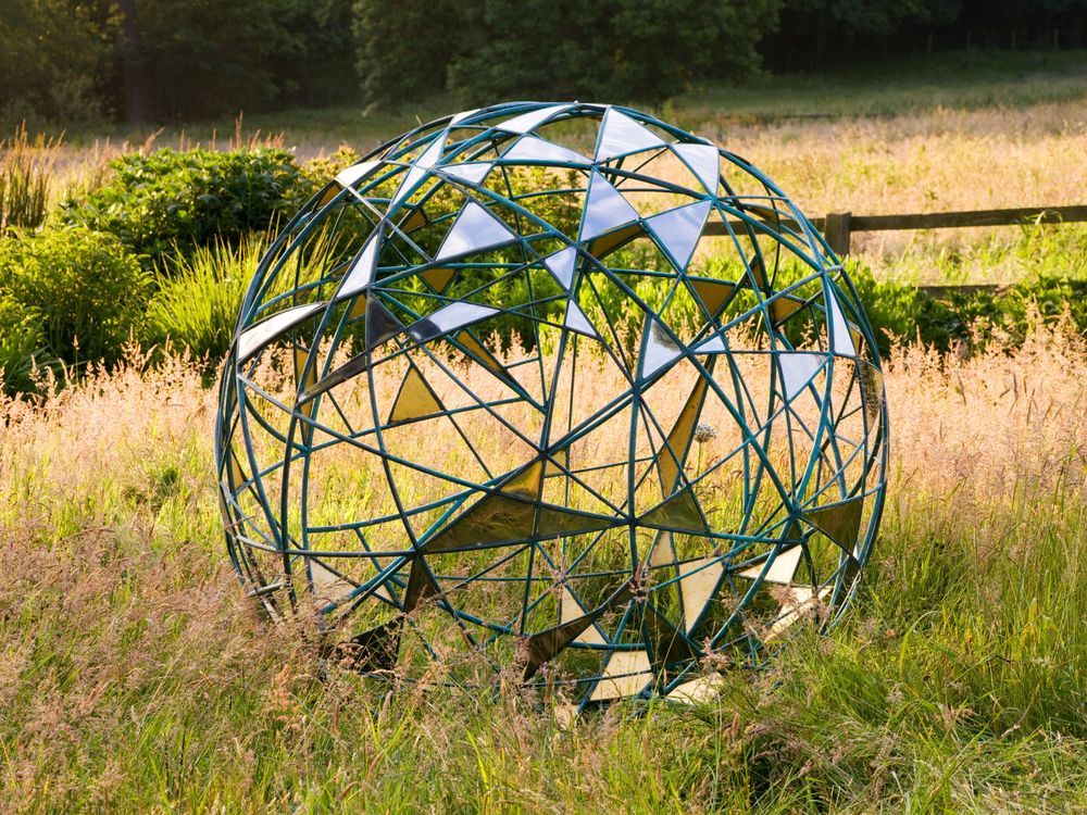 A metal sphere with mirrored panels sits in tall grass in a sunlit, naturalistic landscape.