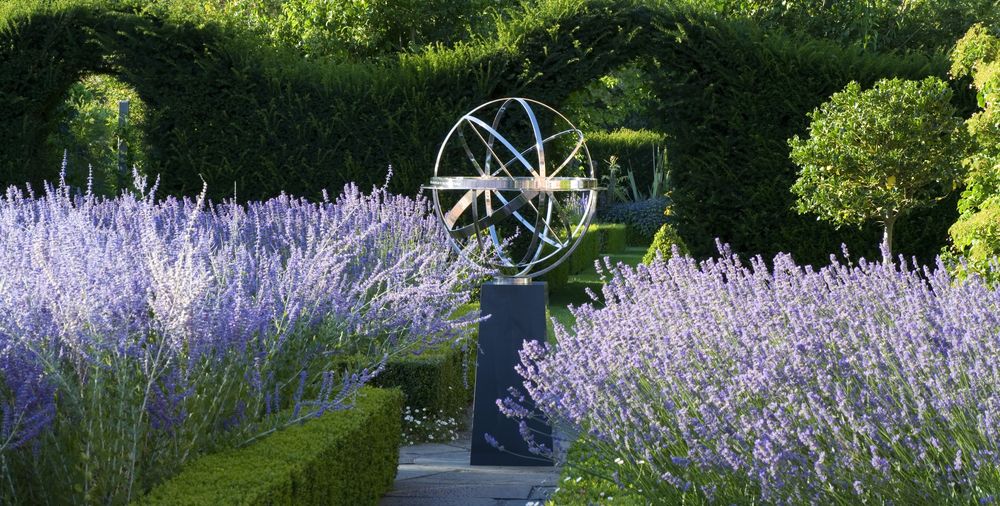 A metal armillary sphere sculpture stands amid lush lavender and green hedges in a garden.