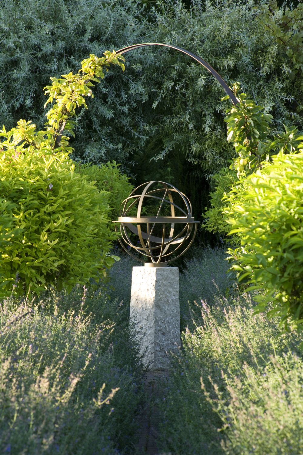 A metal armillary sphere on a stone pedestal, framed by green shrubs and an arched trellis in a garden.