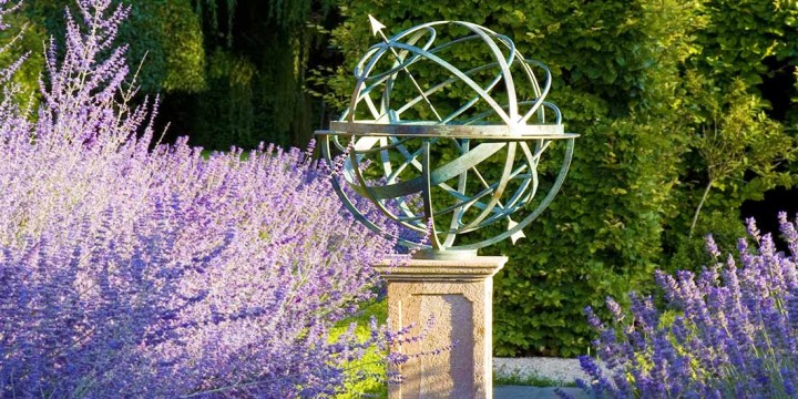 A bronze armillary sphere sundial with a gentle verdigris finish stands on a stone pedestal amid dense purple lavender flowers, lush green foliage in the background, all bathed in bright sunlight.