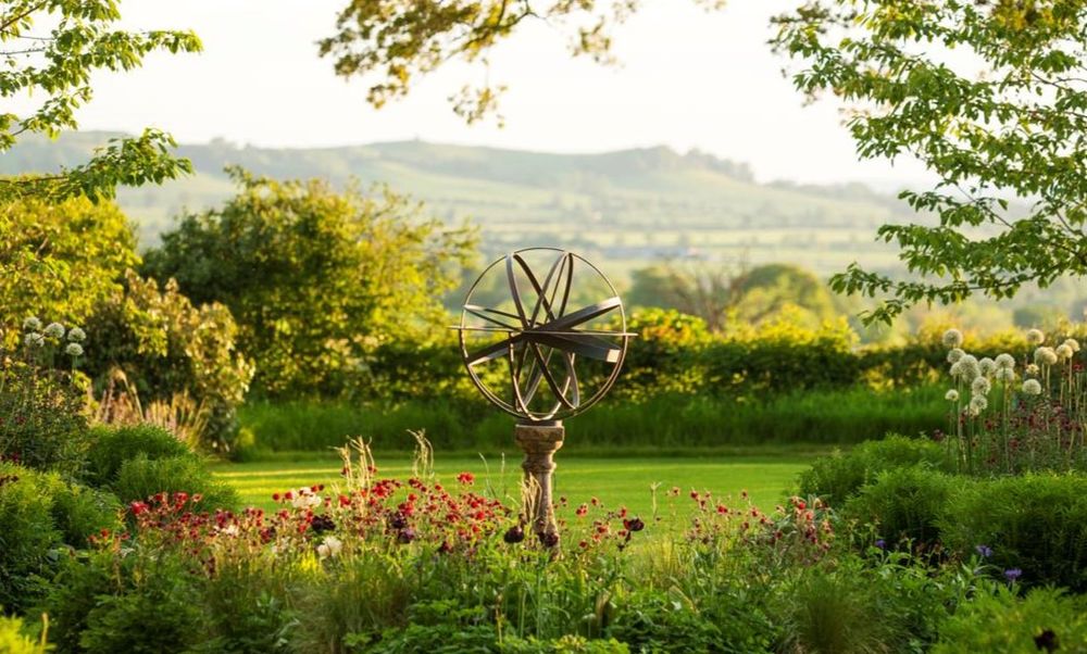 A brass armillary sphere stands in a lush garden with hills and fields in the background.