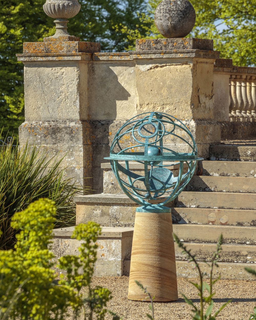 A turquoise metal armillary sphere sculpture on a pedestal in a sunlit garden by stone steps.