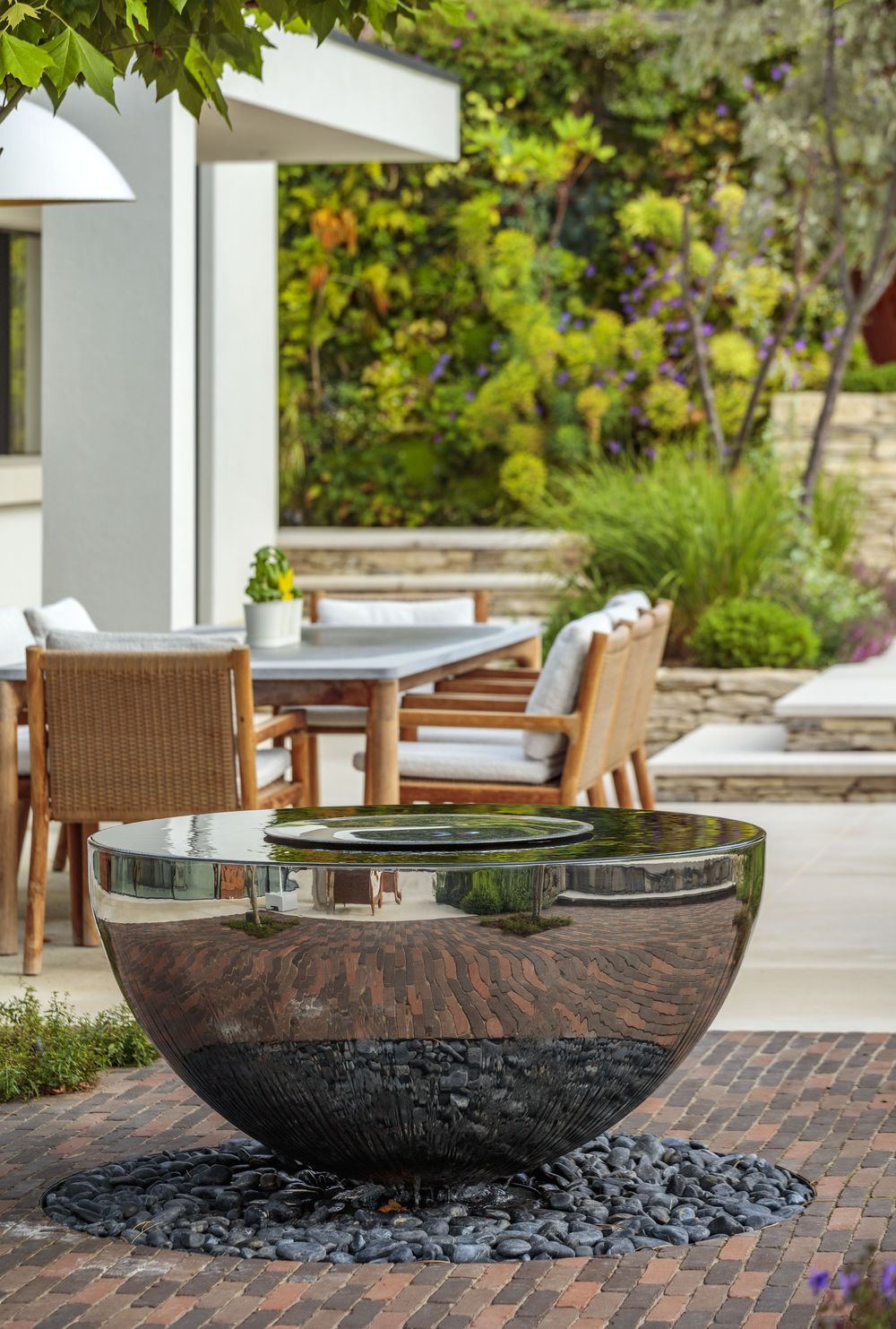 Large chalice stainless steel water bowl fountain on brick patio, near outdoor dining table and lush garden greenery.