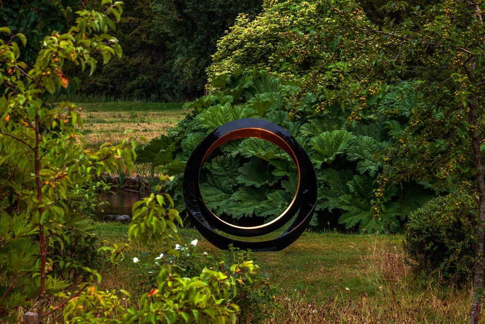 A large black eclipse sculpture stands on grass, surrounded by lush green trees and plants in a garden.
