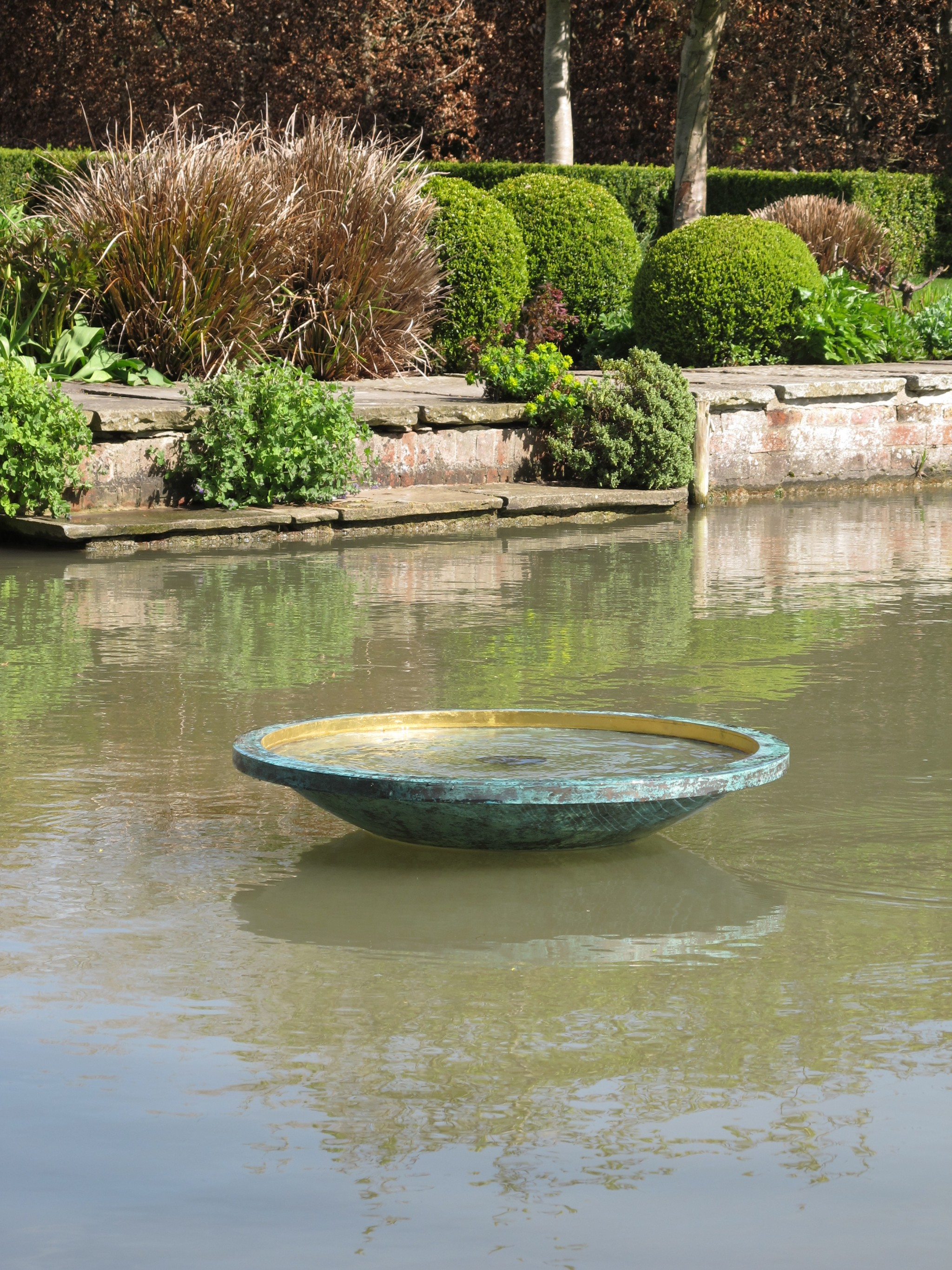 A large, shallow metal bowl—ekho in bronze and gold leaf—floats on a pond amid trimmed garden bushes.