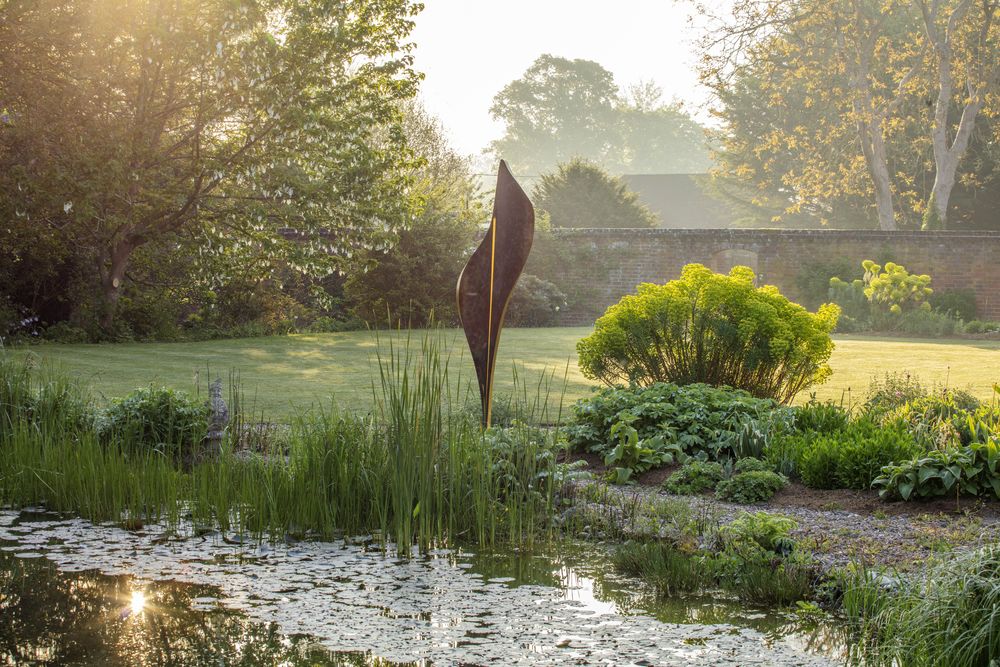 A tranquil garden scene at sunrise features a tall Flo Contemporary Abstracts sculpture made from bronze finish and gold leaf near lush green plants by a pond. Sunlight gently illuminates the landscape, creating a peaceful atmosphere.