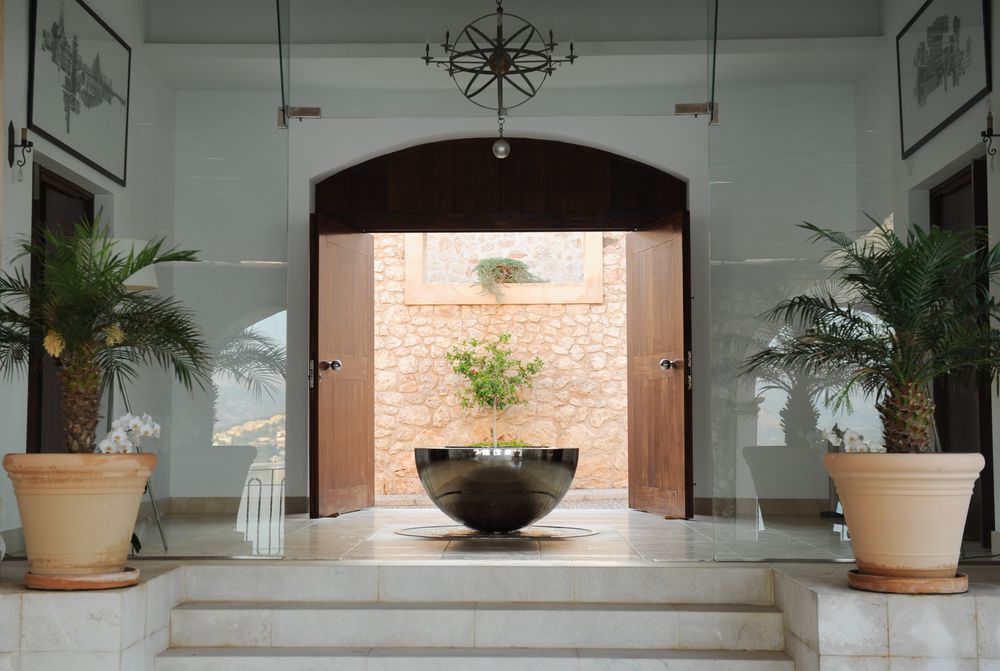 Modern entrance with a large metal bowl, potted palm plants, and a floor sculpture; open wooden doors reveal a stone wall.