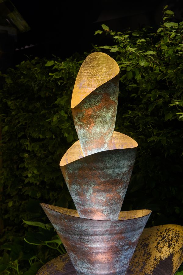 A Florio copper garden contemporary sculpture, this sculptural fountain features three irregular, bowl-like forms stacked vertically, each with mottled green and rust-colored patina, illuminated at night against dense green foliage.
