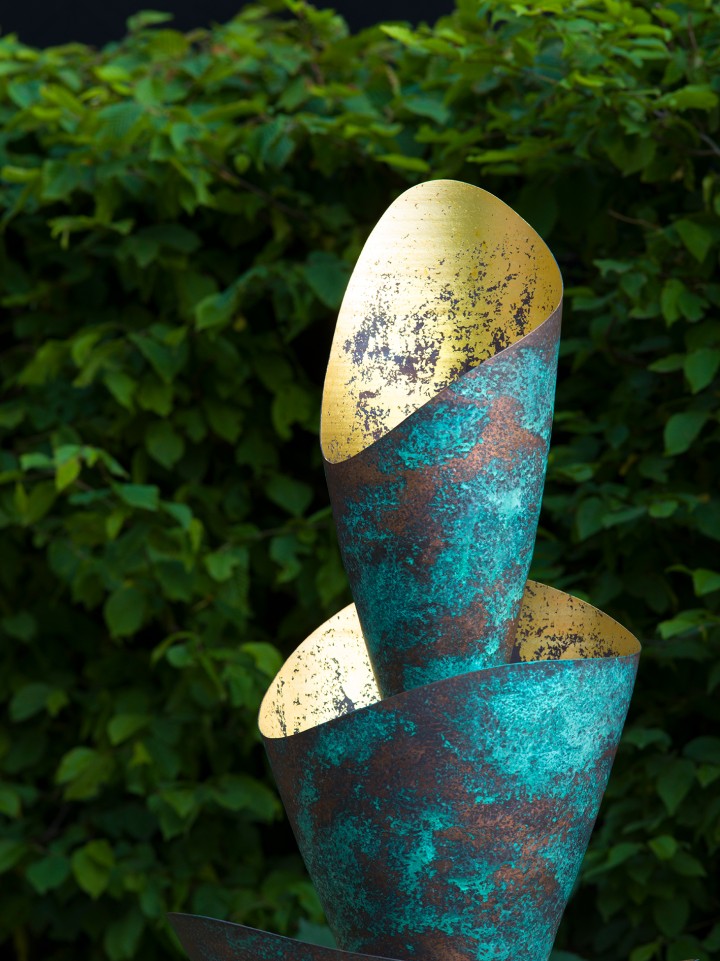 A modern florio sculpture made from verdigris copper patina and 24-carat gold leaf, with overlapping curled metal sheets—teal and rusted outside, shiny mottled gold inside—stands before a lush backdrop of dense green leaves.