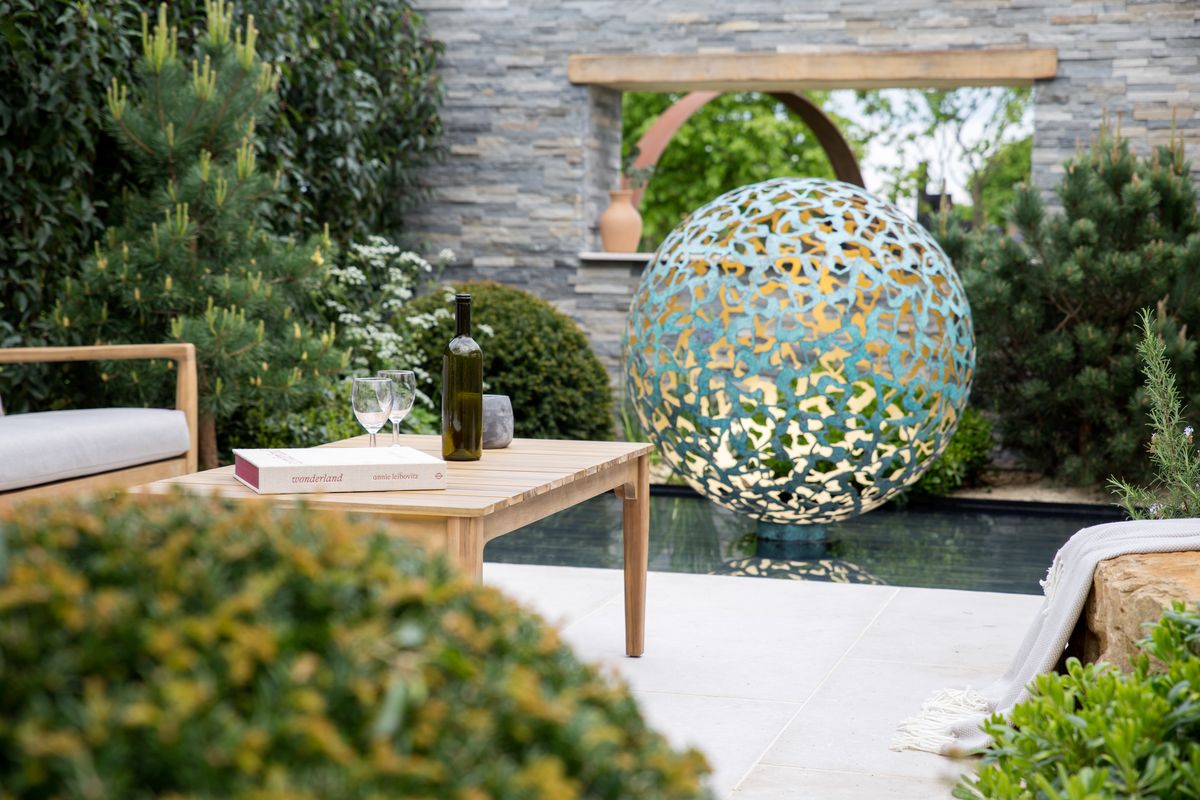 Modern garden with a wooden table, wine glasses, and a metal sphere, perfect for courtyard garden ideas by the swimming pool.
