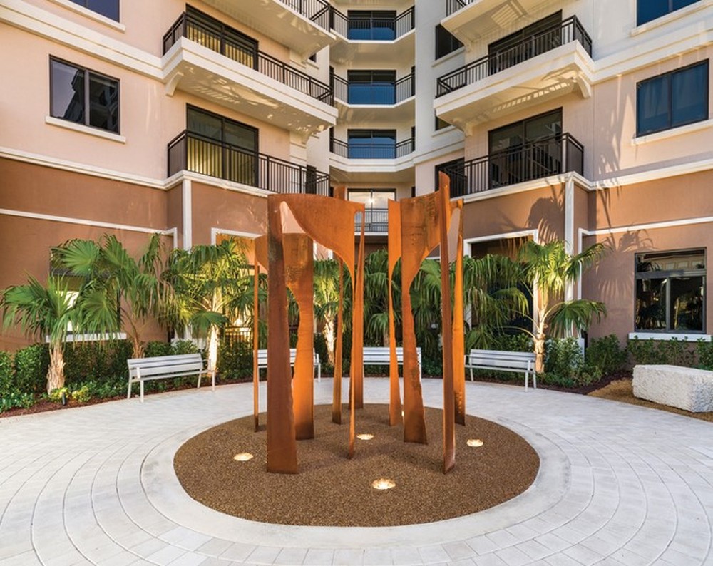 A modern courtyard features a circular area with a glide-inspired contemporary abstract sculpture in rust-colored metal, surrounded by gravel. Palm trees, benches, and manicured shrubs line the space against a multi-story building with balconies and large windows.